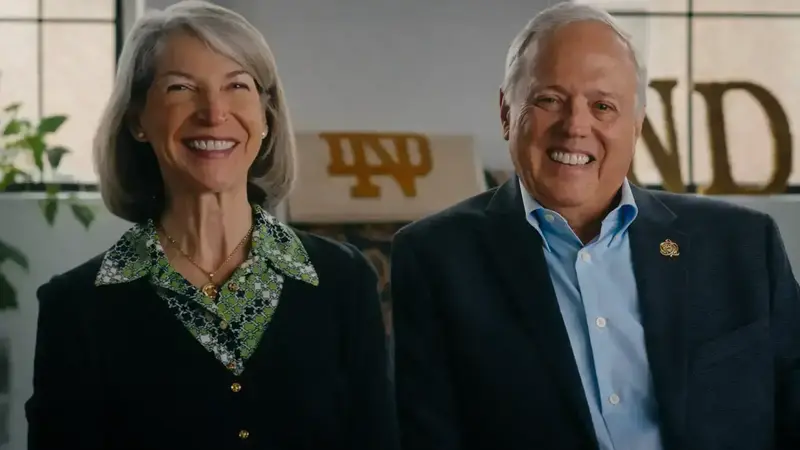 A woman and man sit next to each other and smile broadly. She wears a green patterned shirt; he wears a blue shirt with a lapel pin. A large ND logo is behind them.
