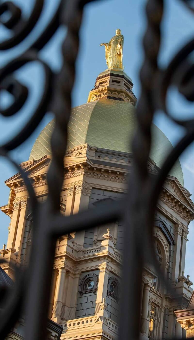 Golden Dome of Notre Dame's Main Building and Our Lady statue, framed by ornate dark iron gate against blue sky.